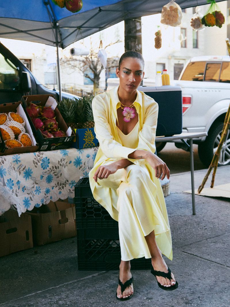 A woman in a yellow outfit poses at an outdoor fruit stand, seated on a black crate, with tropical fruits and a white truck nearby.