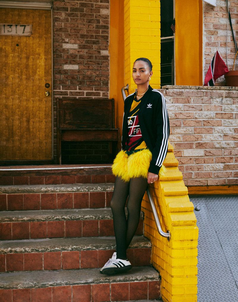 A stylish woman in an Adidas jacket and yellow skirt poses on brick steps in front of a yellow door, showcasing a fashion-forward look.