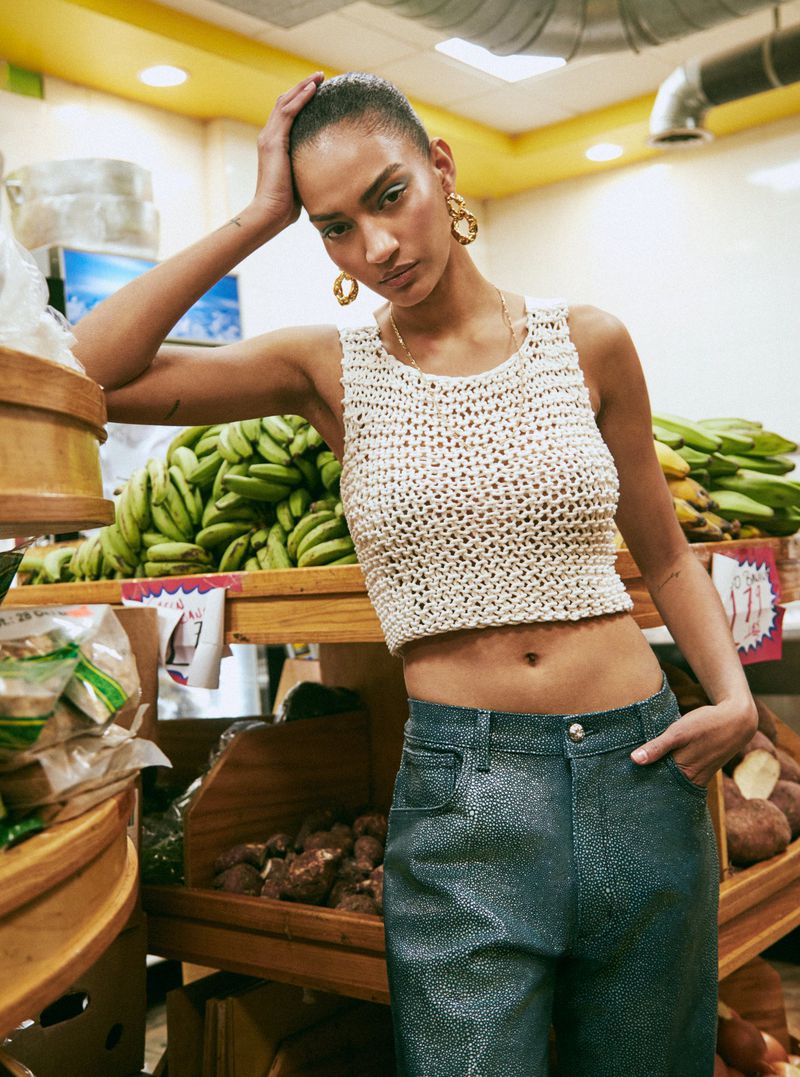 Adult female posing in a light-colored knitted tank top and sparkling blue pants in a grocery store setting surrounded by produce.