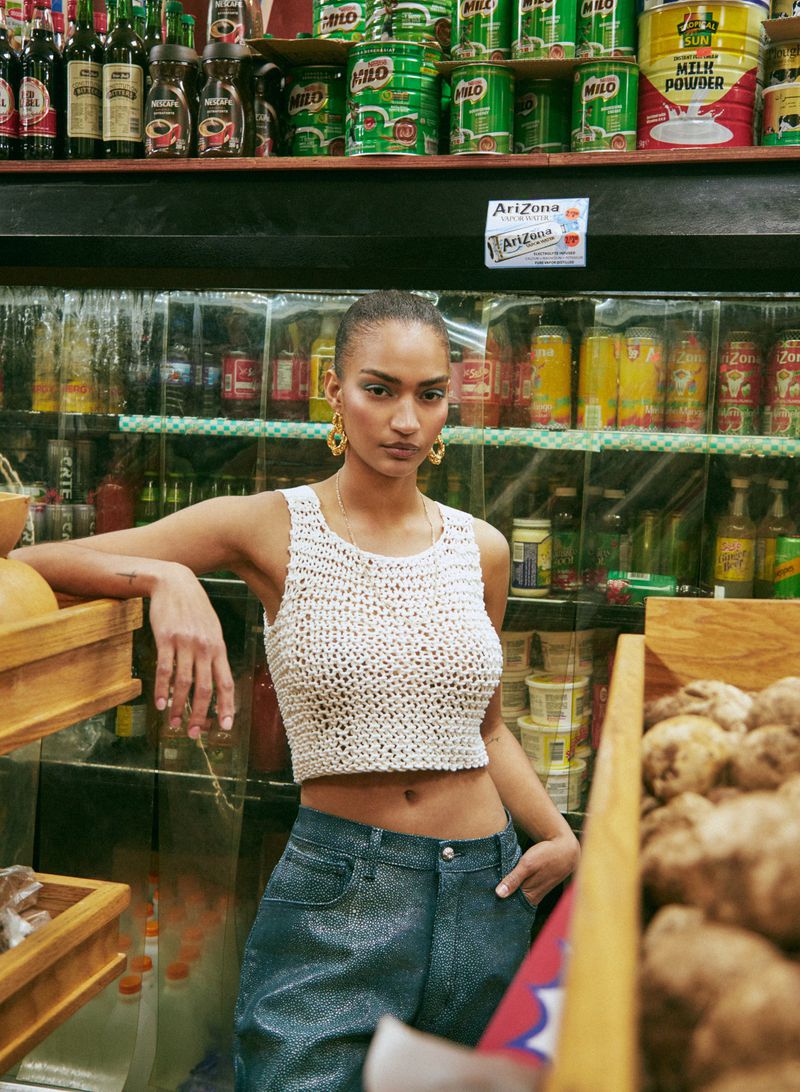 Woman poses in a knit crop top and blue jeans in a store with shelves of food, wearing large hoop earrings and a serious expression.