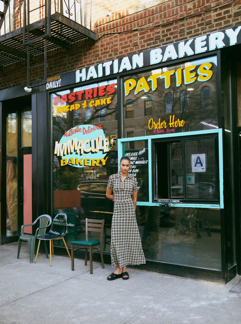A woman stands in front of Immaculate Bakery wearing a plaid dress and sandals on an outdoor city street setting with chairs.