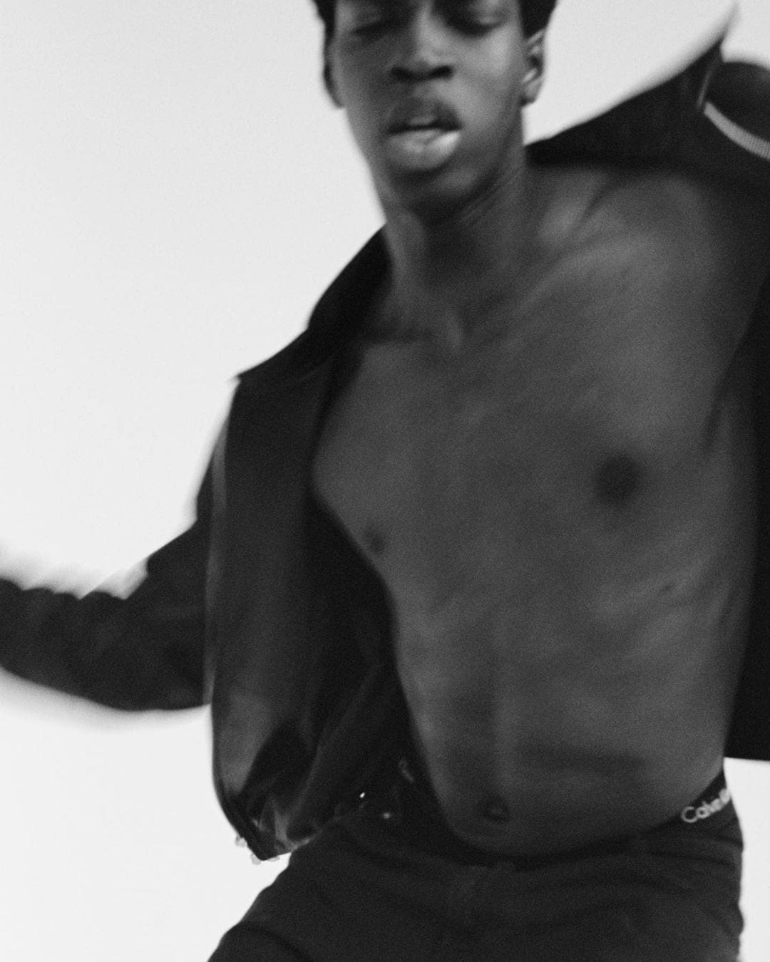 Black and white image of a shirtless man with motion blur in a dark jacket against a plain background.