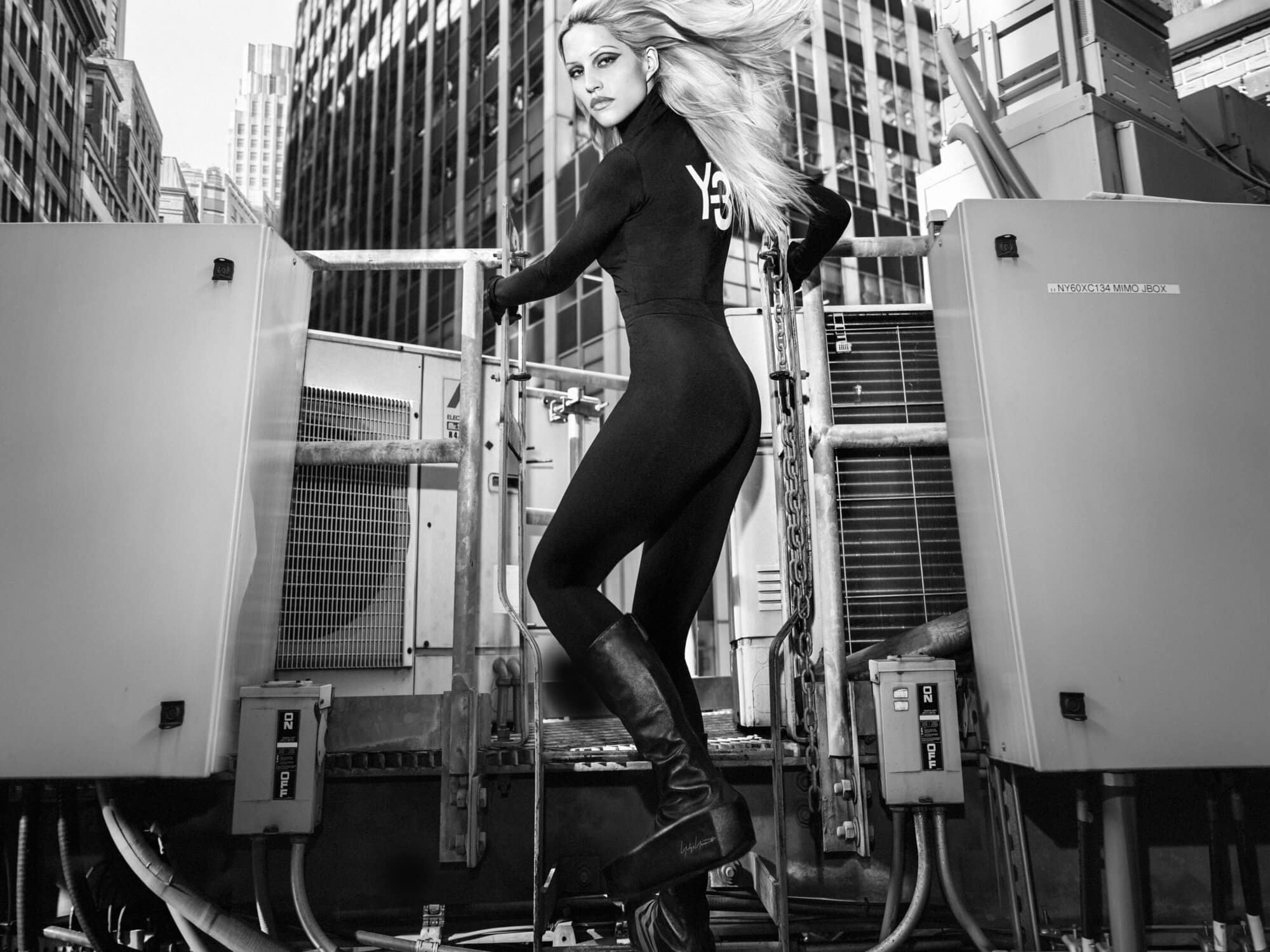 Black and white portrait of Gabbriette wearing a black Y-3 bodysuit and tall boots on an industrial rooftop setting.