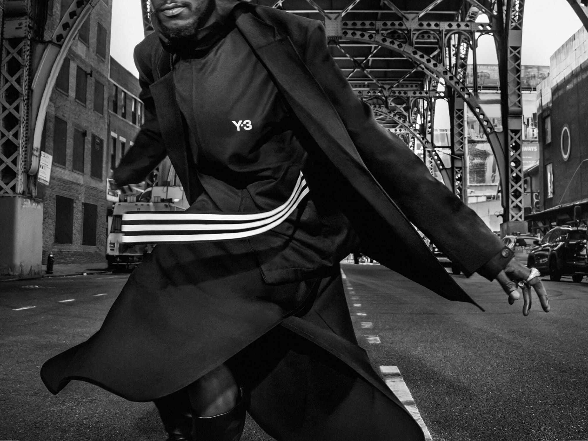 Black and white editorial photograph of a model in motion wearing a Y-3 black coat on an urban street under an elevated railway.