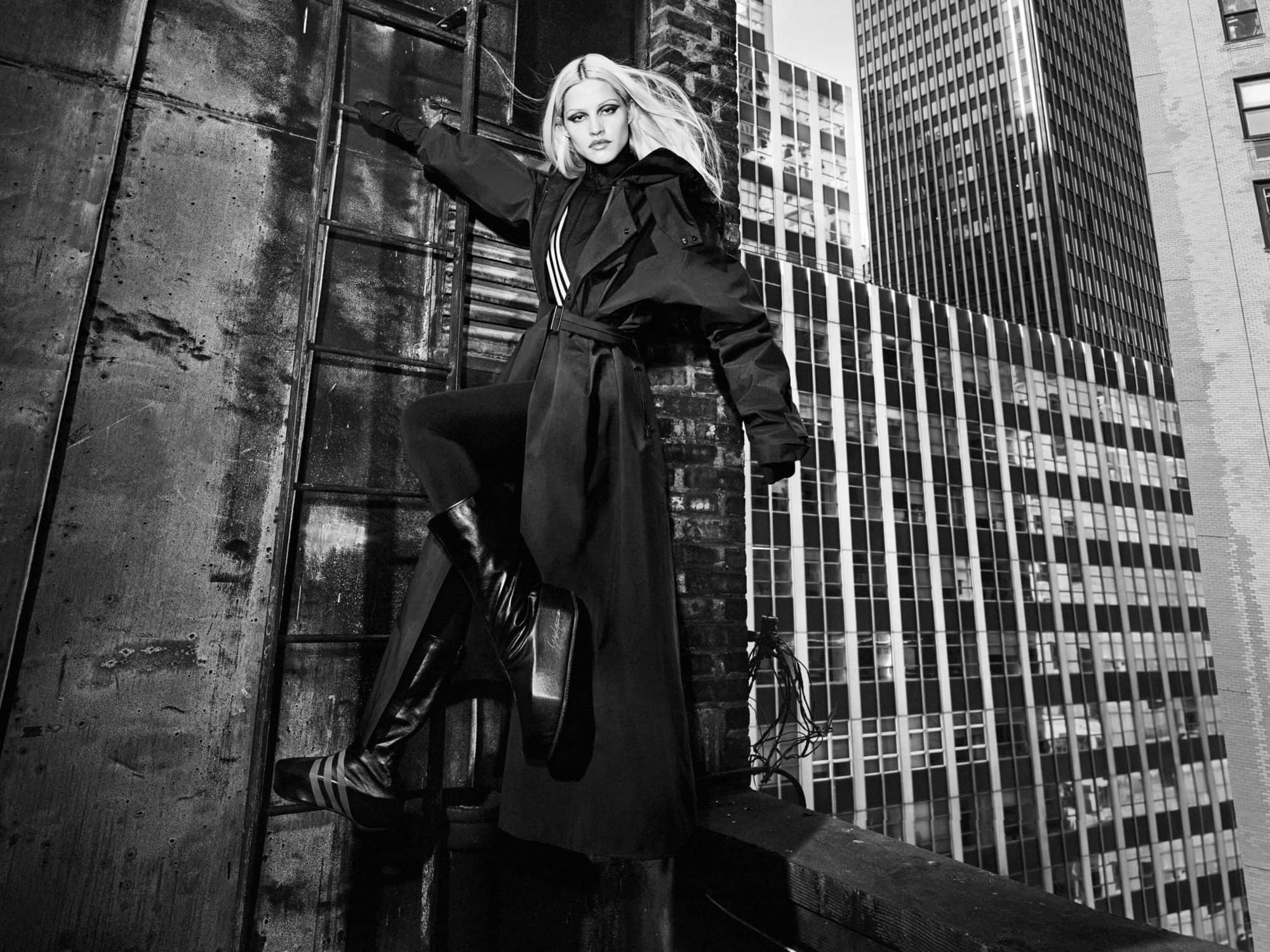 Model Gabbriette wearing a dark coat and boots climbing an outdoor ladder against a backdrop of tall skyscrapers in New York