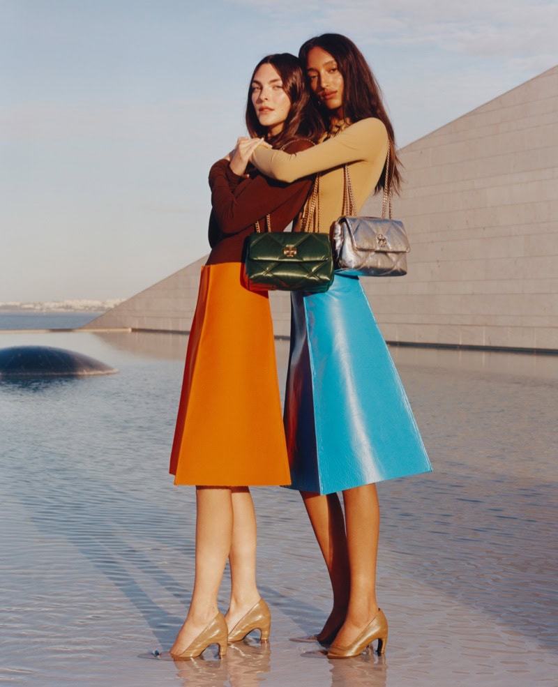 Two models embracing outdoors near a reflective pool wearing colorful midi skirts and carrying quilted shoulder bags