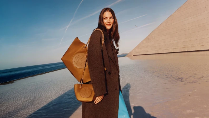 Model in long dark coat with tan chain-strap bags against modern architecture and ocean backdrop for Tory Burch Fall 2024 campaign
