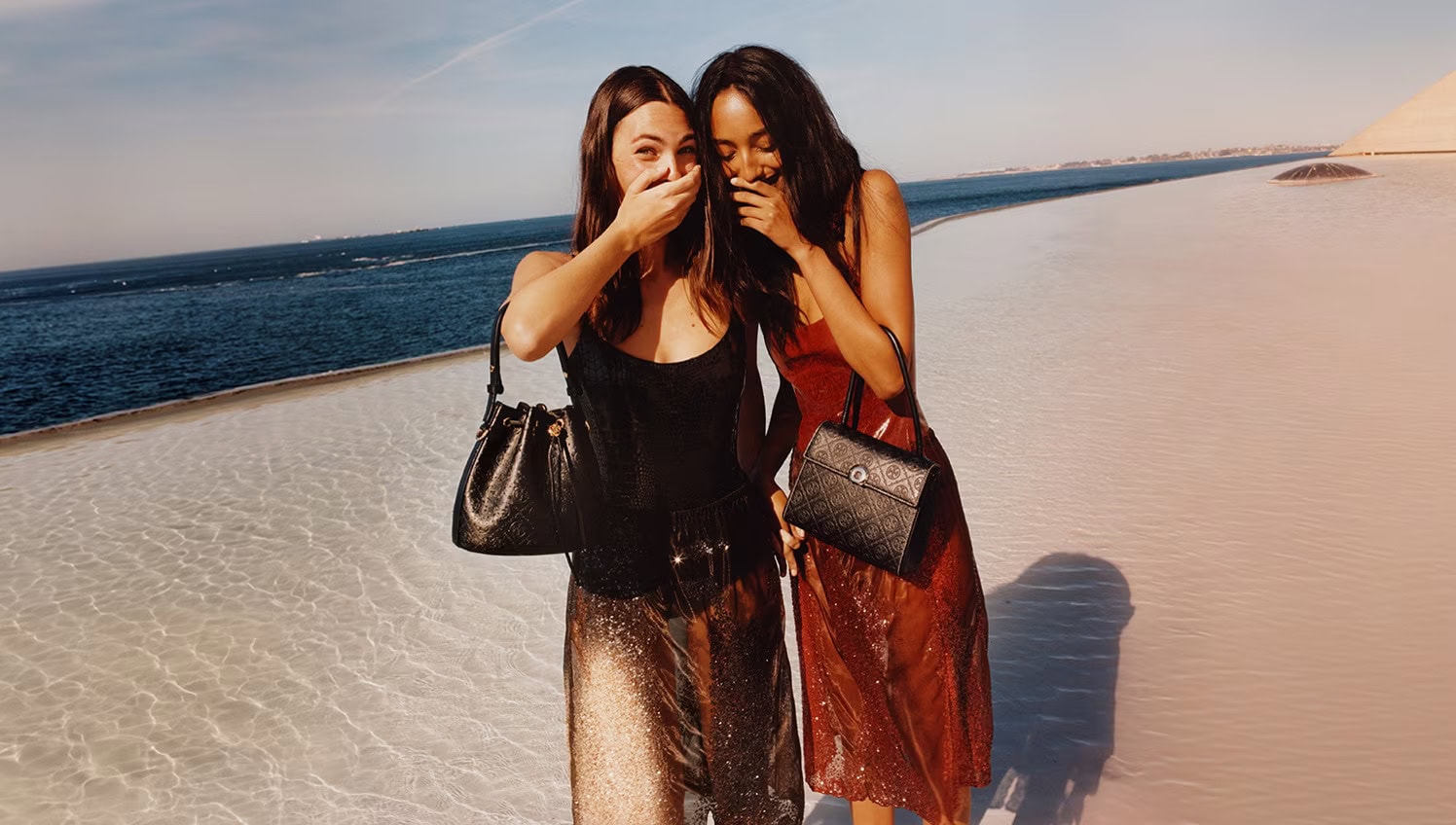 Two models laughing while holding designer handbags by the coast for Tory Burch Fall 2024 ad campaign