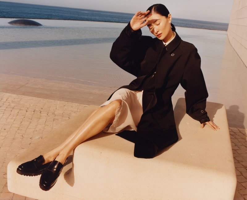 Model wearing a black coat and light skirt seated outdoors near water for Tory Burch