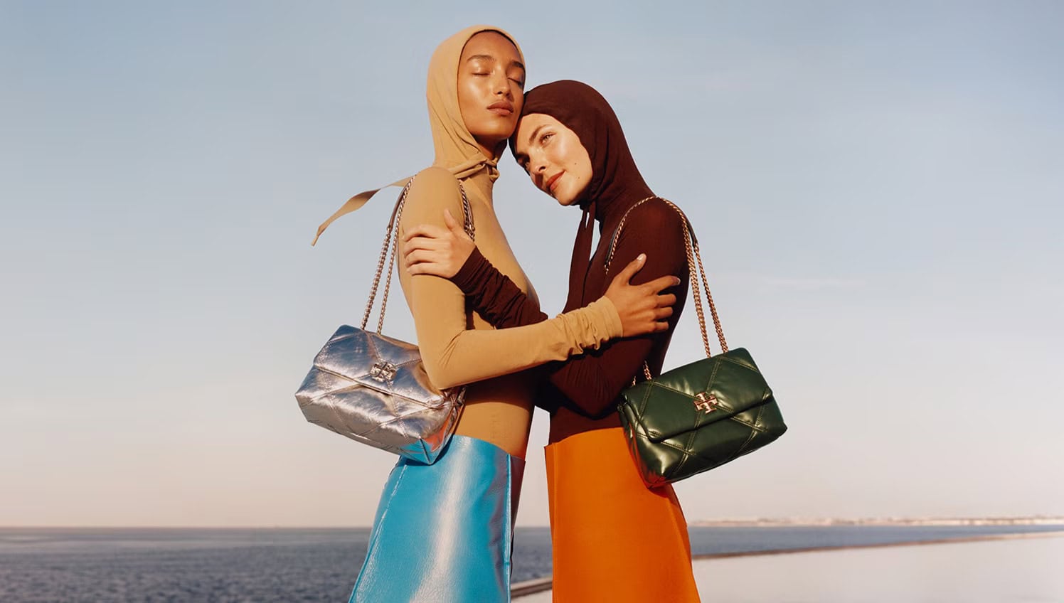 Two models wearing fitted hooded tops and quilted chain-strap handbags against an open horizon.