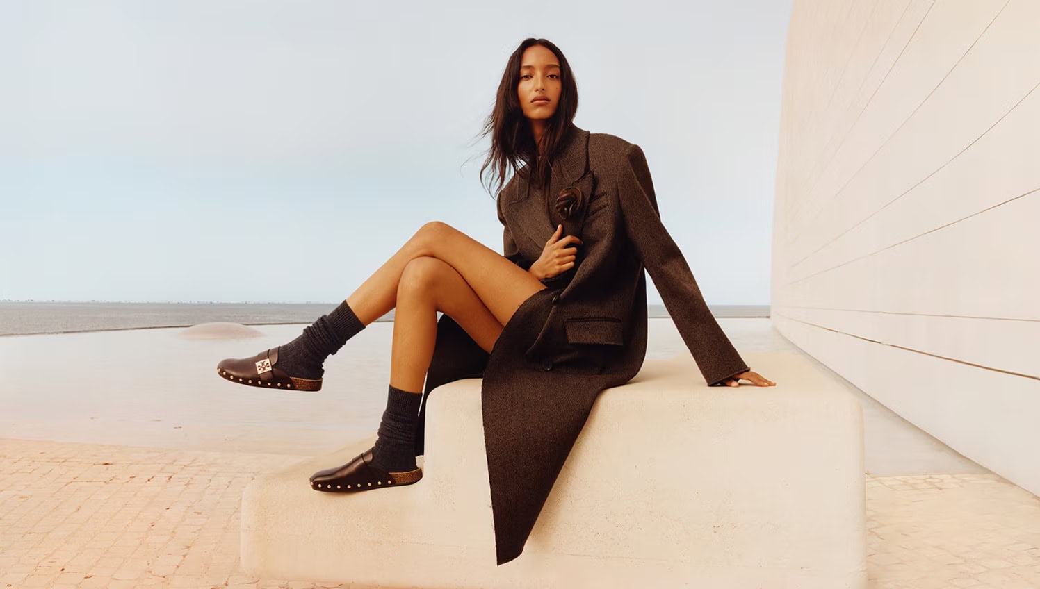 Model seated on concrete ledge outdoors wearing oversized dark blazer coat, crew socks, and studded clogs with ocean background.