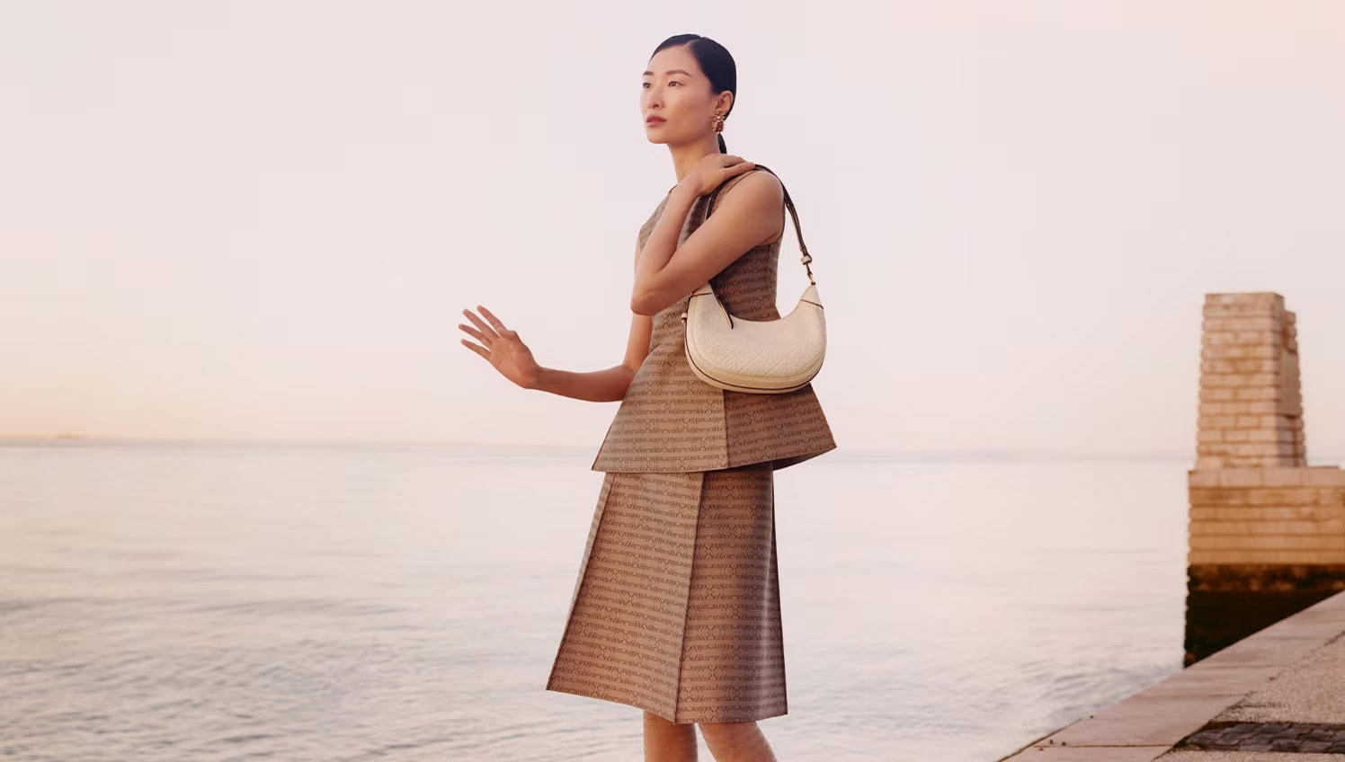 Model in patterned brown peplum dress wearing white shoulder bag beside calm water at sunset