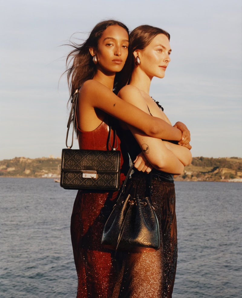 Two female models posing outdoors by the water holding black leather handbags for Tory Burch Fall 2024 campaign