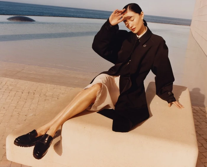 Model wearing a black coat and light skirt seated outdoors near water for Tory Burch