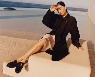 Model wearing a black coat and light skirt seated outdoors near water for Tory Burch