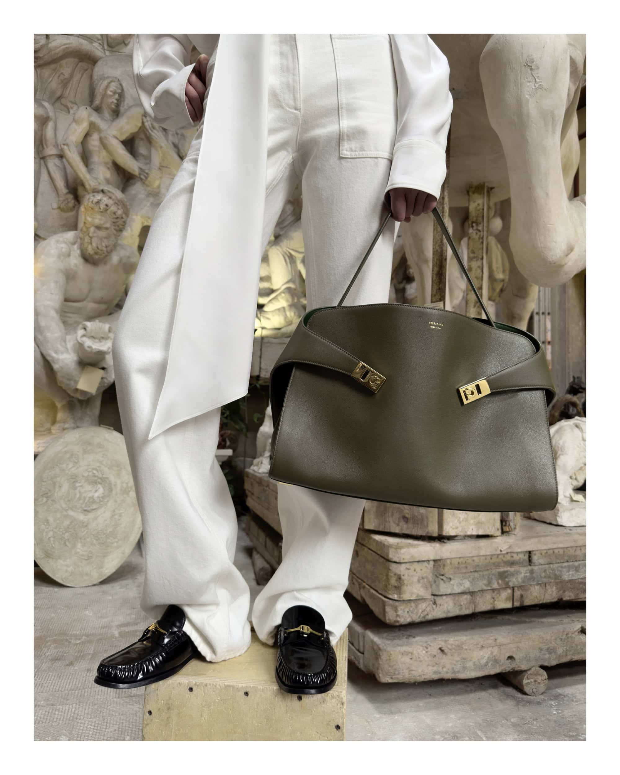 Person in white trousers and black loafers holding a large olive green leather handbag against a sculptural museum background
