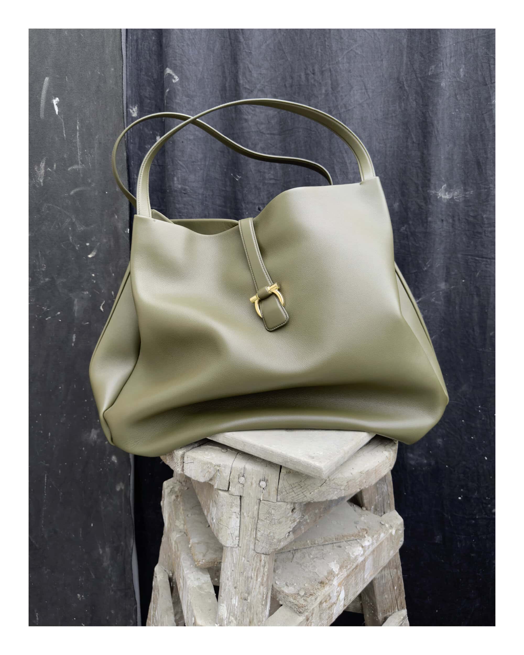 Olive green leather handbag with gold clasp resting on a rustic wooden stool against a dark backdrop