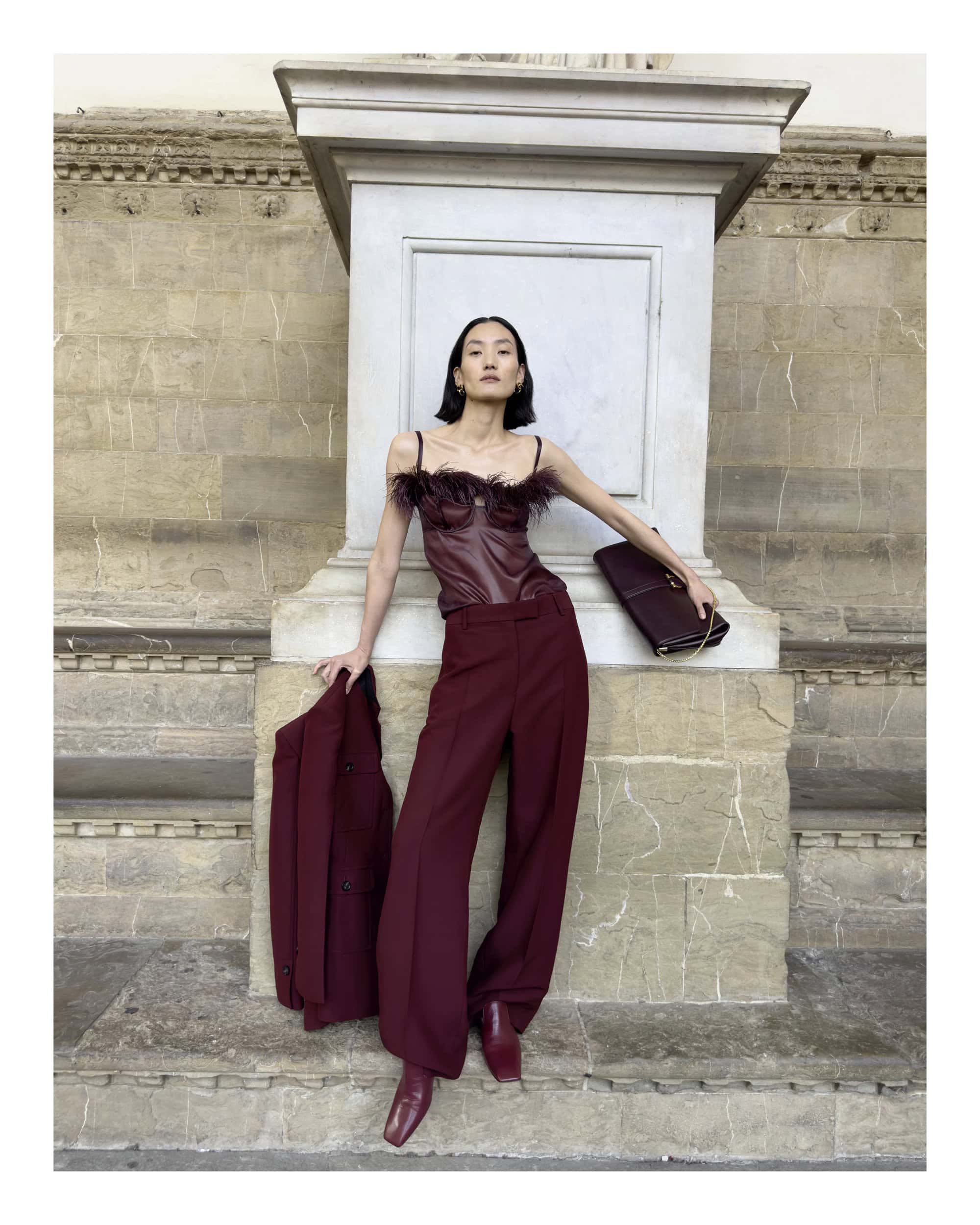 Model wearing a burgundy leather corset top and matching trousers posing against a stone architectural monument