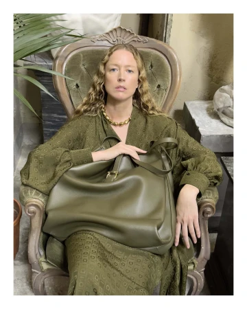 Woman in an olive green patterned dress seated in an ornate chair holding a large matching leather shoulder bag