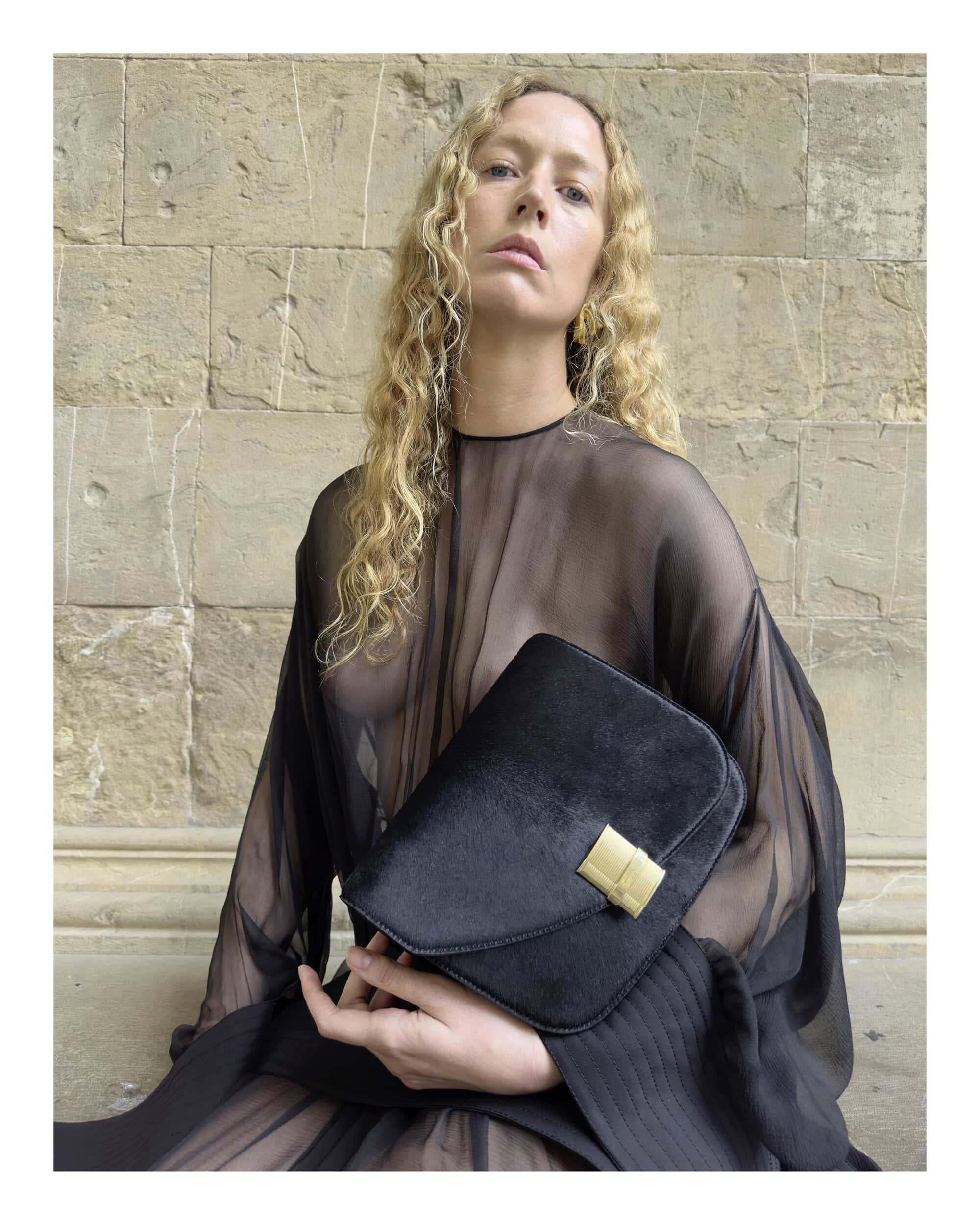 Woman with long blonde curly hair wearing a sheer black dress holding a black handbag in front of a stone wall.