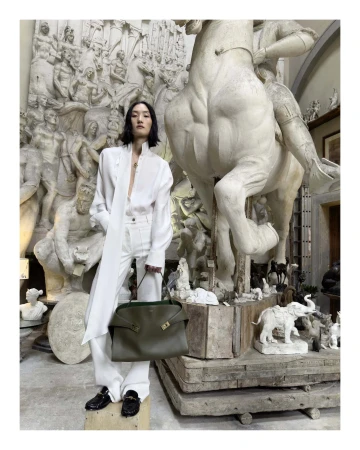 Model in white shirt and pants holding olive green handbag standing in museum gallery with large classical sculptures