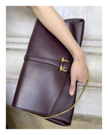 Close-up of a person holding a large dark burgundy leather clutch bag with a gold chain strap against a stone wall.