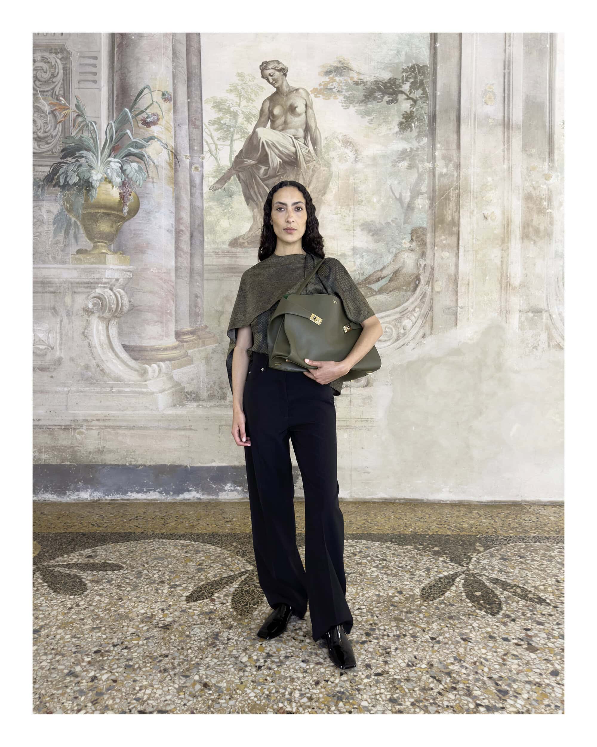 Model wearing a draped top and black trousers standing against a classic fresco wall holding a large olive green handbag.