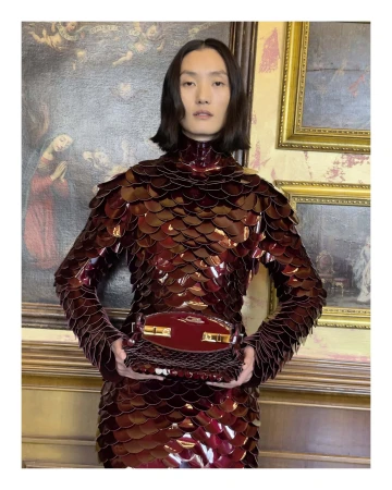 Model wearing a glossy burgundy scaled top holding a matching handbag in front of classical framed paintings in Florence