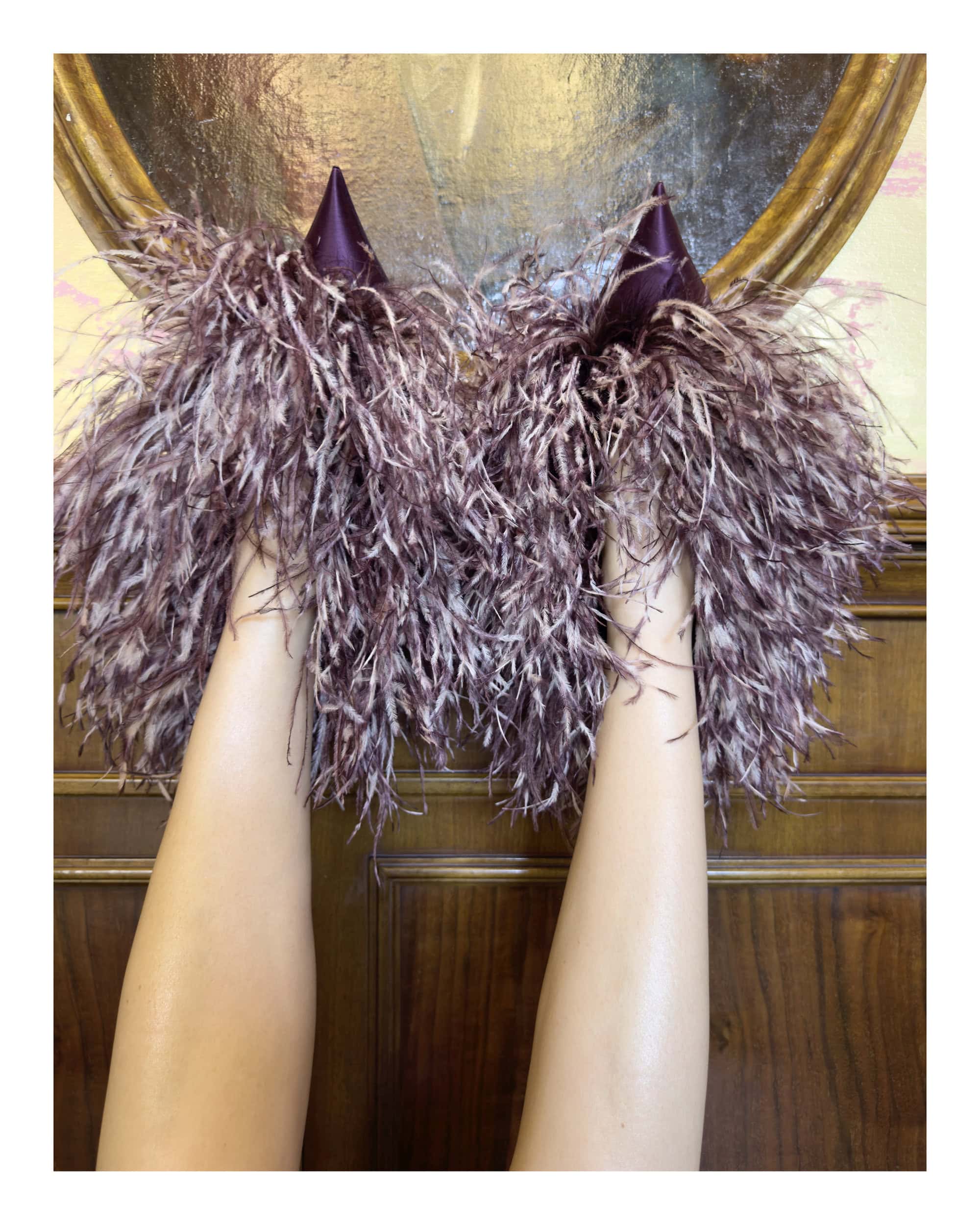 Close-up of legs held up, wearing feathered purple pointy shoes against wooden paneling in Ferragamo 2024 campaign by Juergen Teller