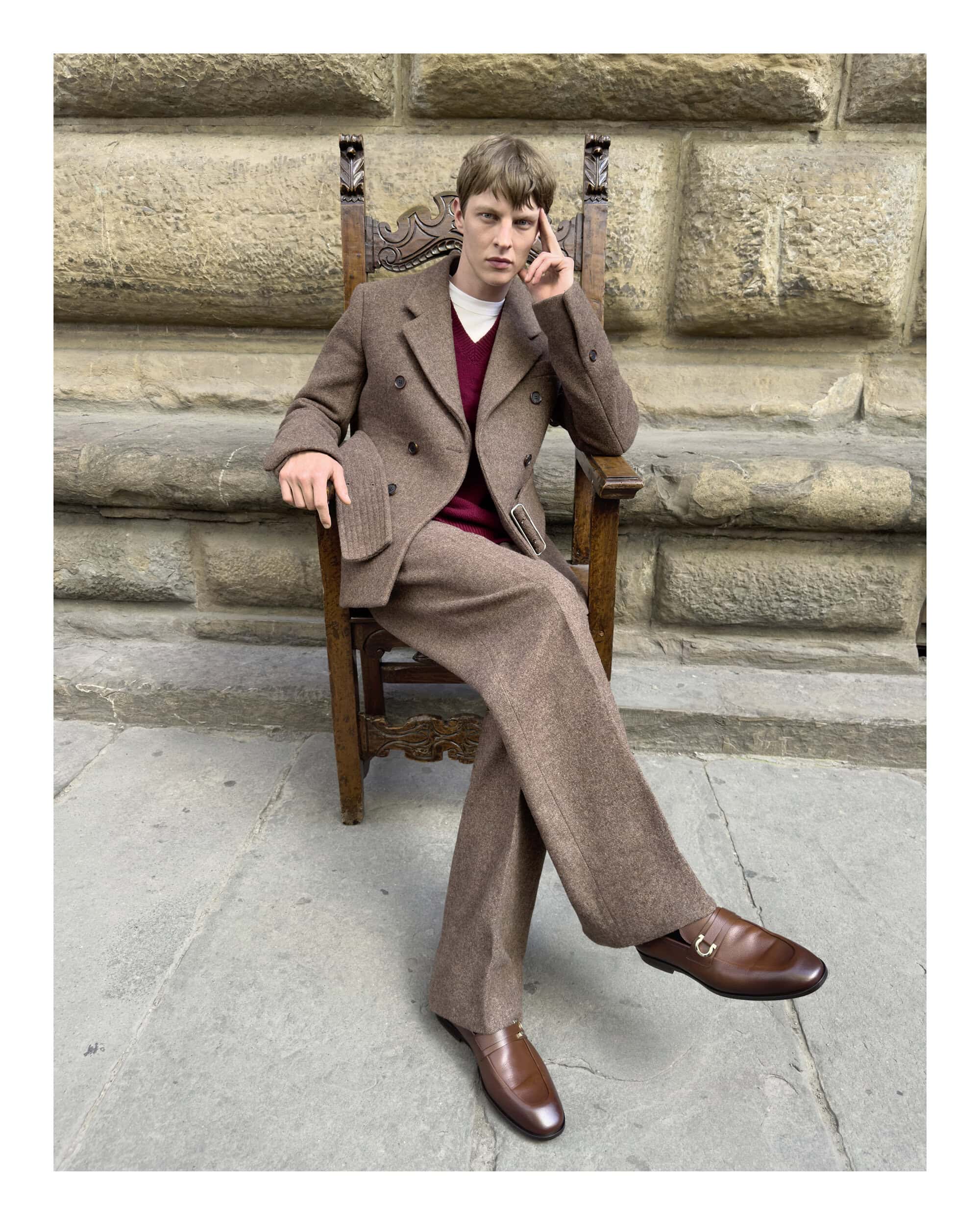 Man in a brown tweed suit sitting on a carved wooden chair outdoors against a rustic stone wall