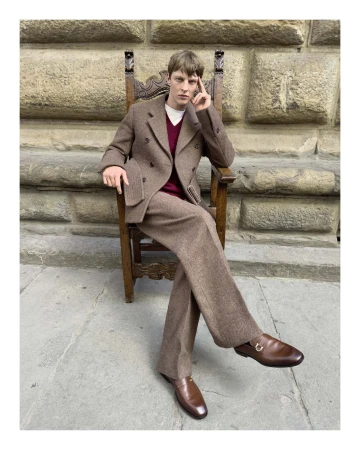 Man in a brown tweed suit sitting on a carved wooden chair outdoors against a rustic stone wall