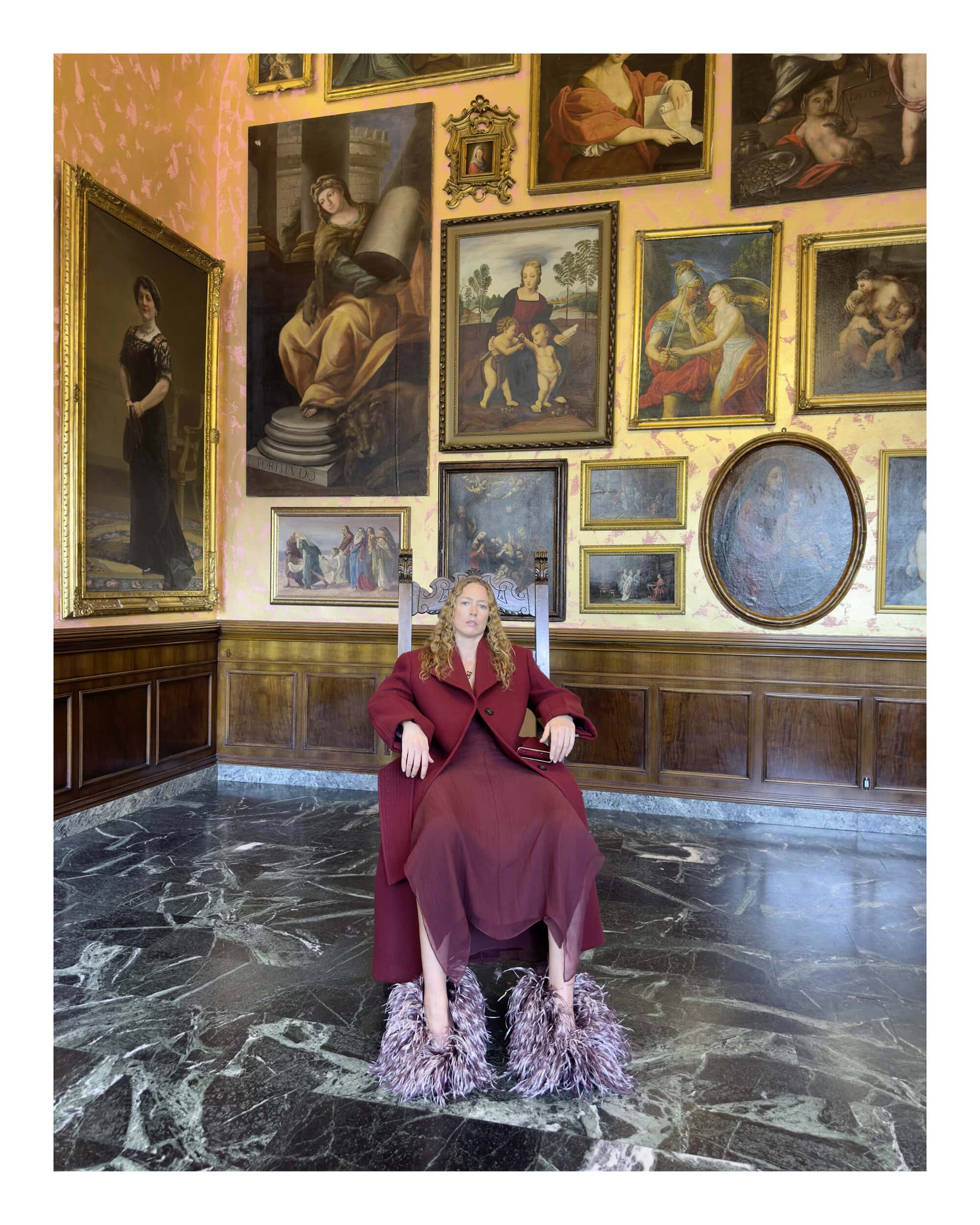 Model seated on a high-backed wooden chair wearing a burgundy coat and fringed shoes inside a gallery filled with paintings