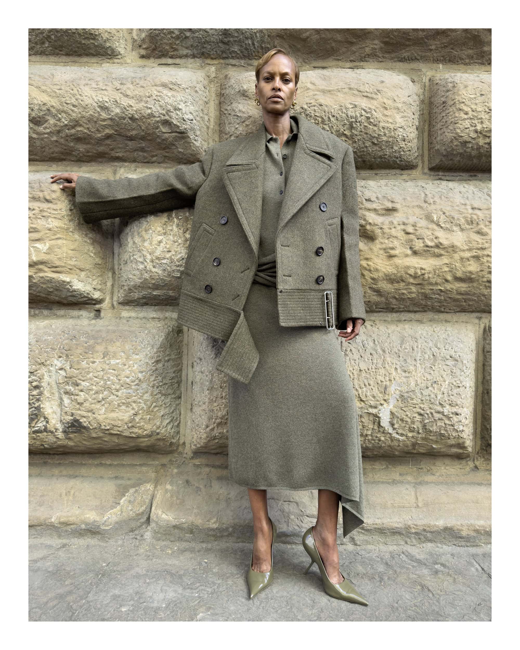 Model standing against a weathered stone wall wearing an olive green coat, matching skirt, and pointed-toe heels.