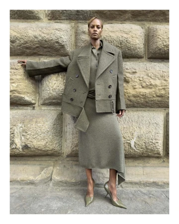 Model standing against a weathered stone wall wearing an olive green coat, matching skirt, and pointed-toe heels.