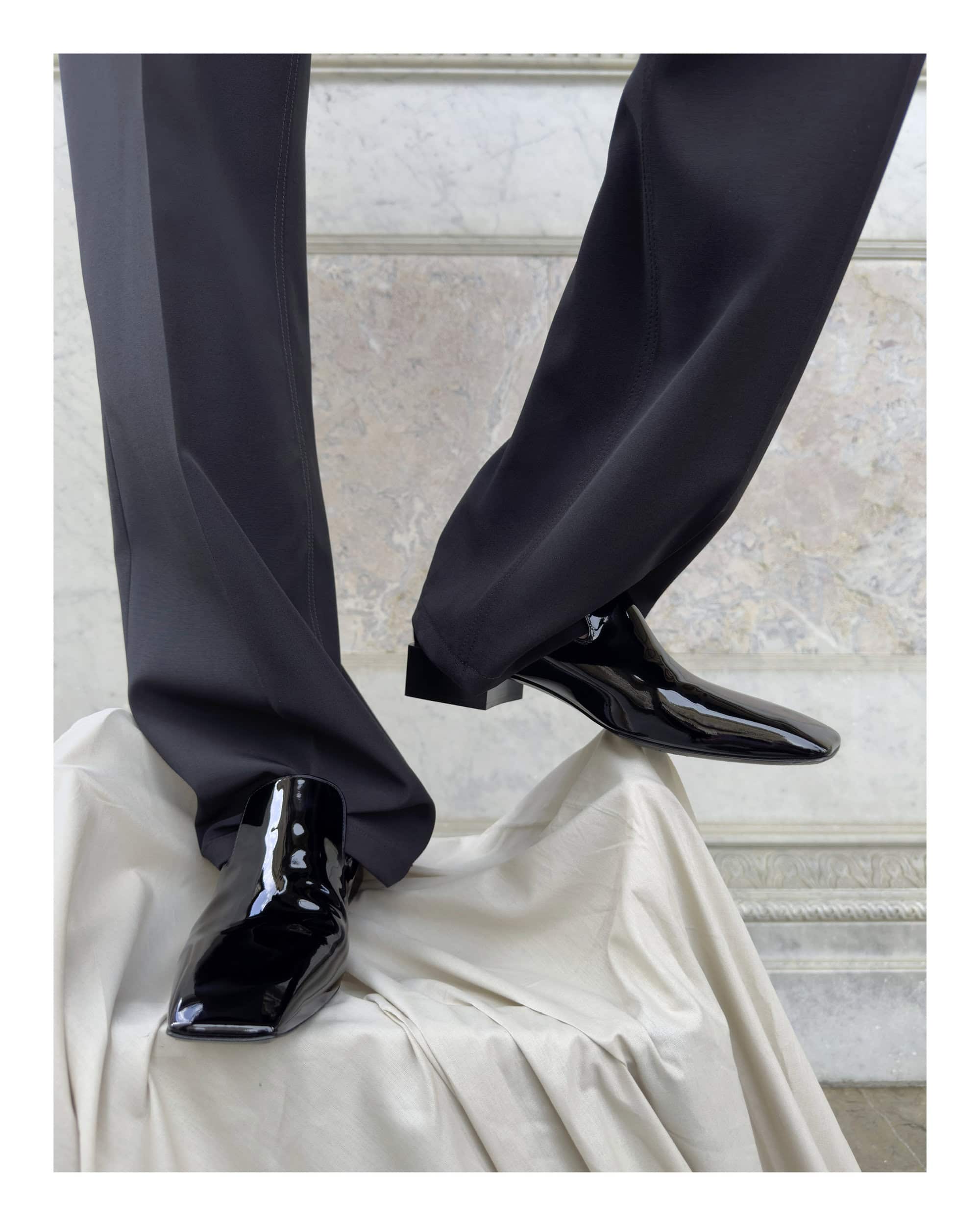 Close-up shot of patent black leather shoes and dark trousers resting on draped white fabric against marble architecture.