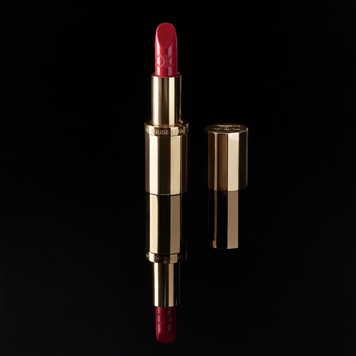 Celine lipstick tube in gold casing with red bullet standing vertically against a solid black reflective background.