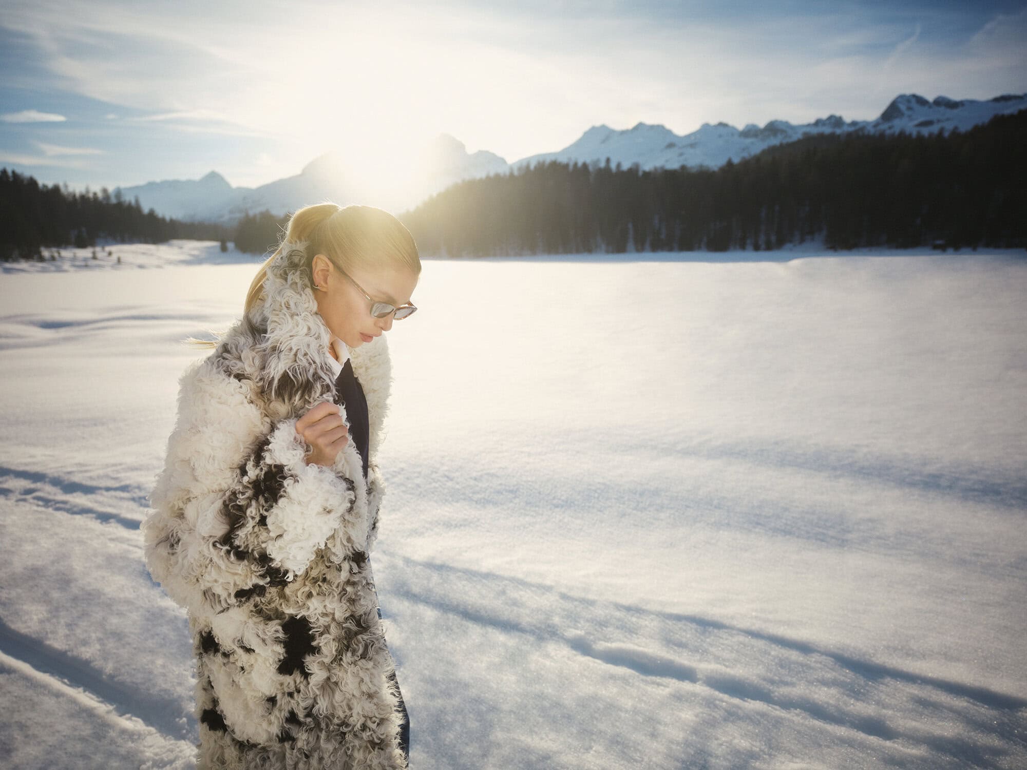Model wearing a fuzzy patterned coat and sunglasses outdoors in a snowy landscape with mountains under a bright sun