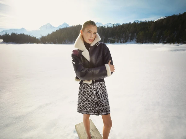 Model standing outdoors on a snowy landscape wearing a dark leather shearling jacket and patterned skirt