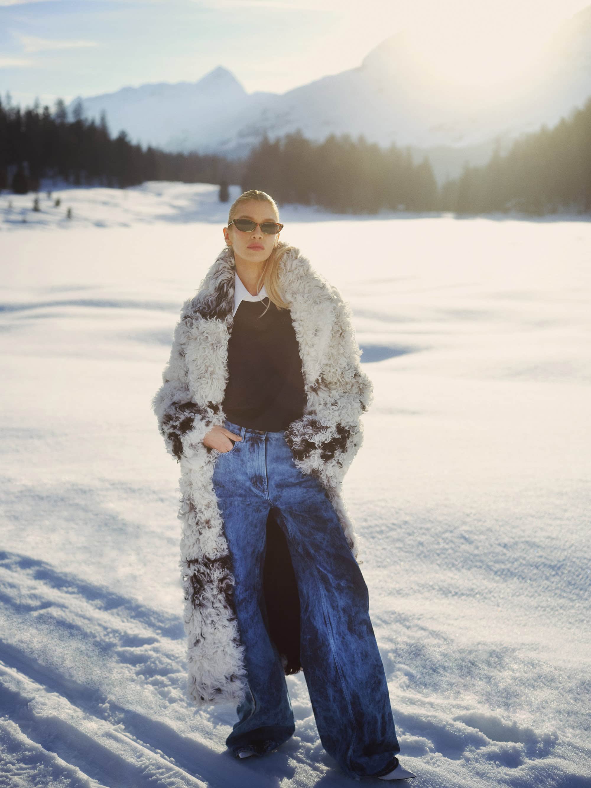 Model in sunglasses wearing a fluffy shearling coat and blue jeans standing on a snowy mountain landscape
