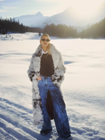 Model in sunglasses wearing a fluffy shearling coat and blue jeans standing on a snowy mountain landscape