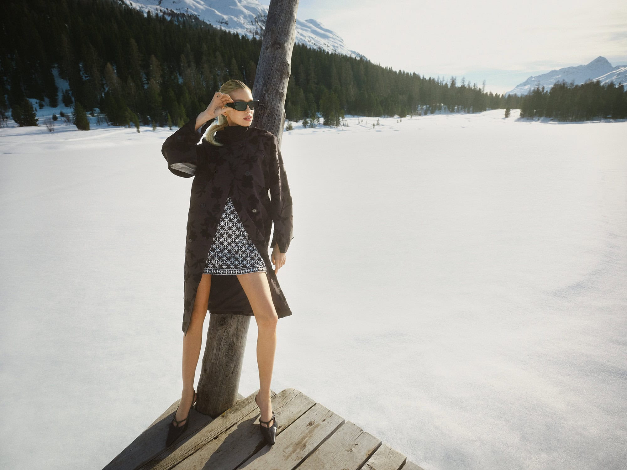 Model standing on a wooden dock in a snowy landscape wearing a dark coat and patterned dress