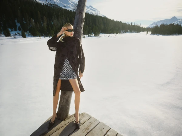 Model standing on a wooden dock in a snowy landscape wearing a dark coat and patterned dress