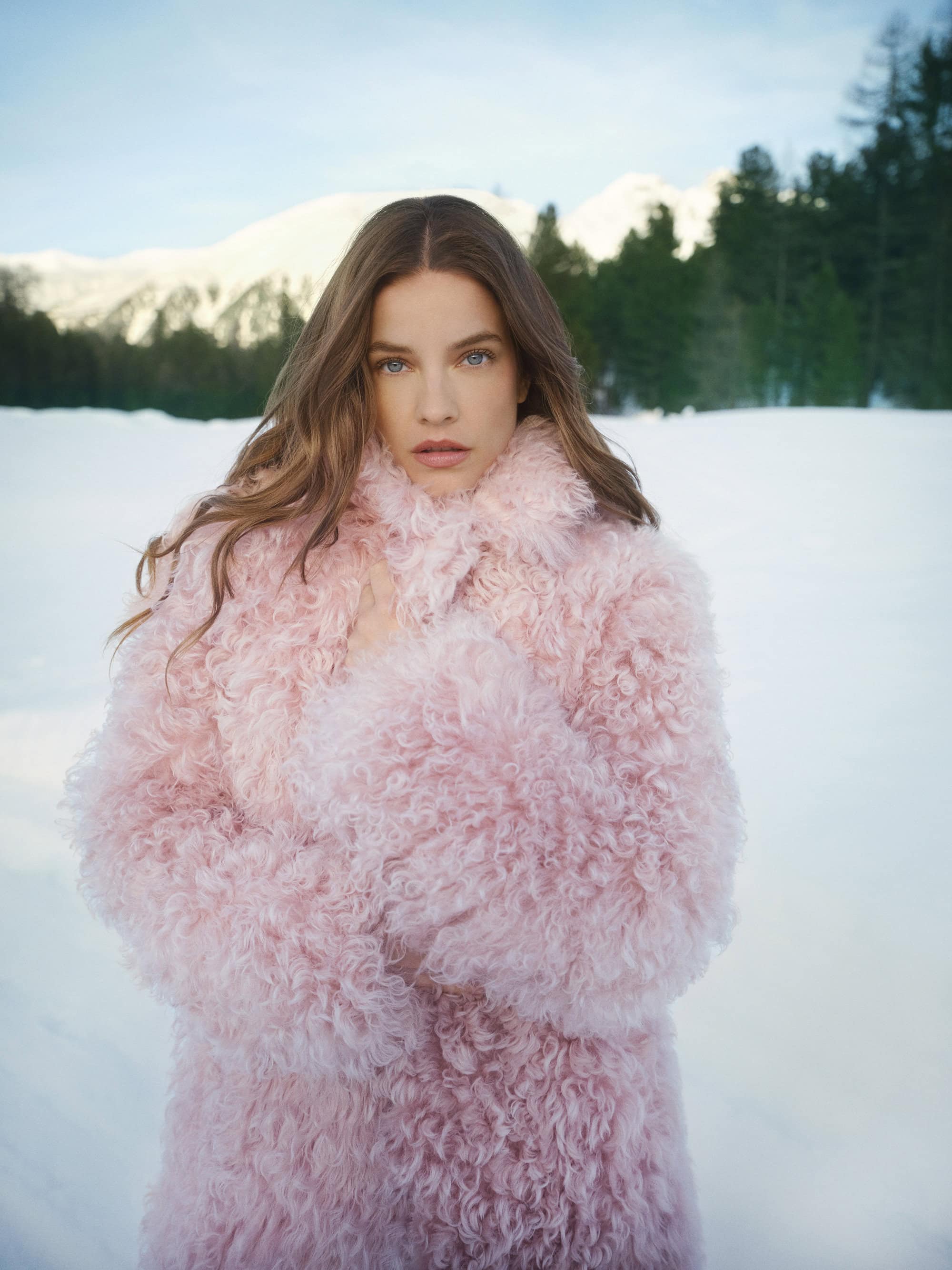 Model Barbara Palvin wearing a fluffy pink coat standing outdoors against a snowy landscape with distant mountains