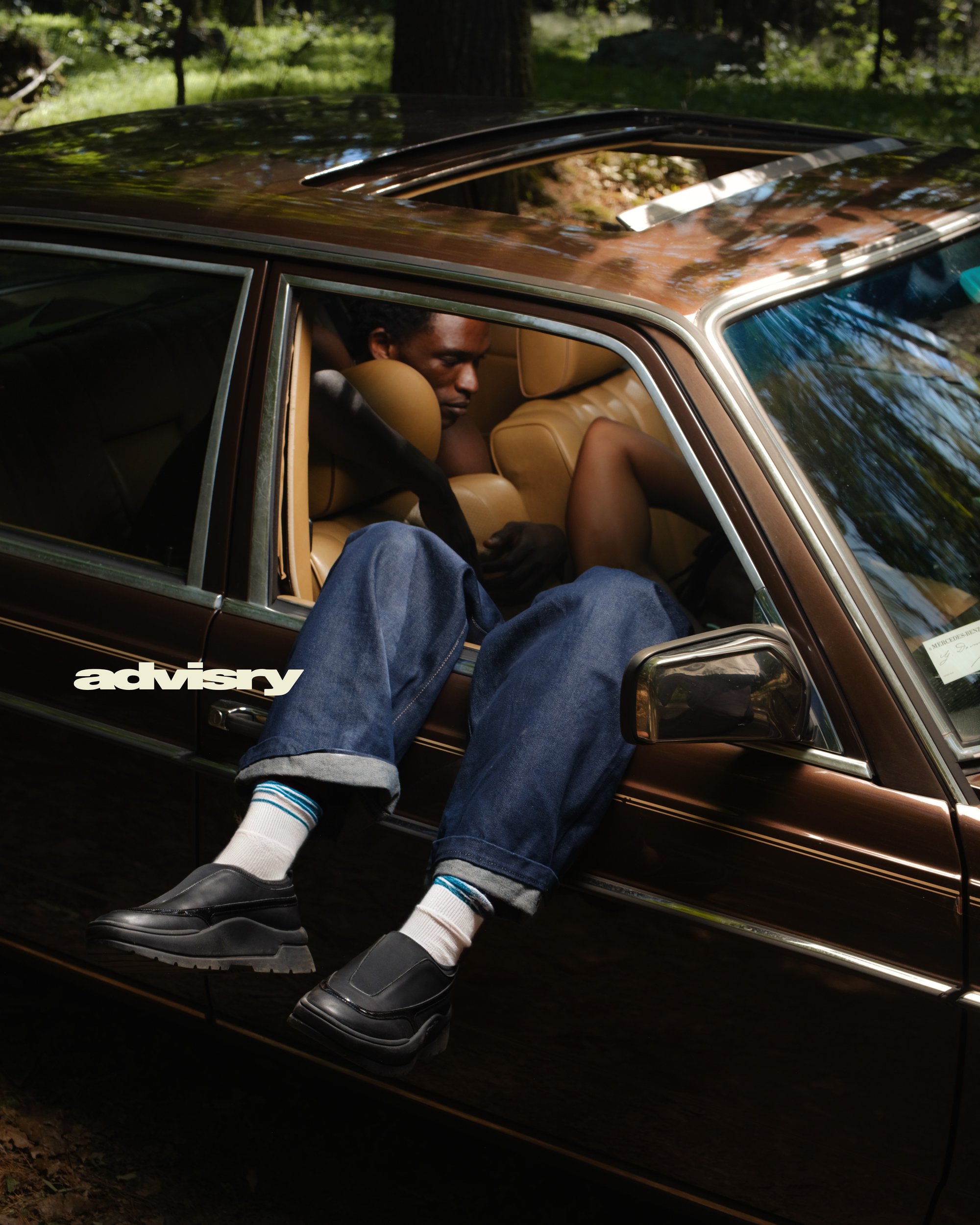 Two Black men seated inside a vintage brown car with legs hanging out of the open window for Advisry Summer 2024 campaign