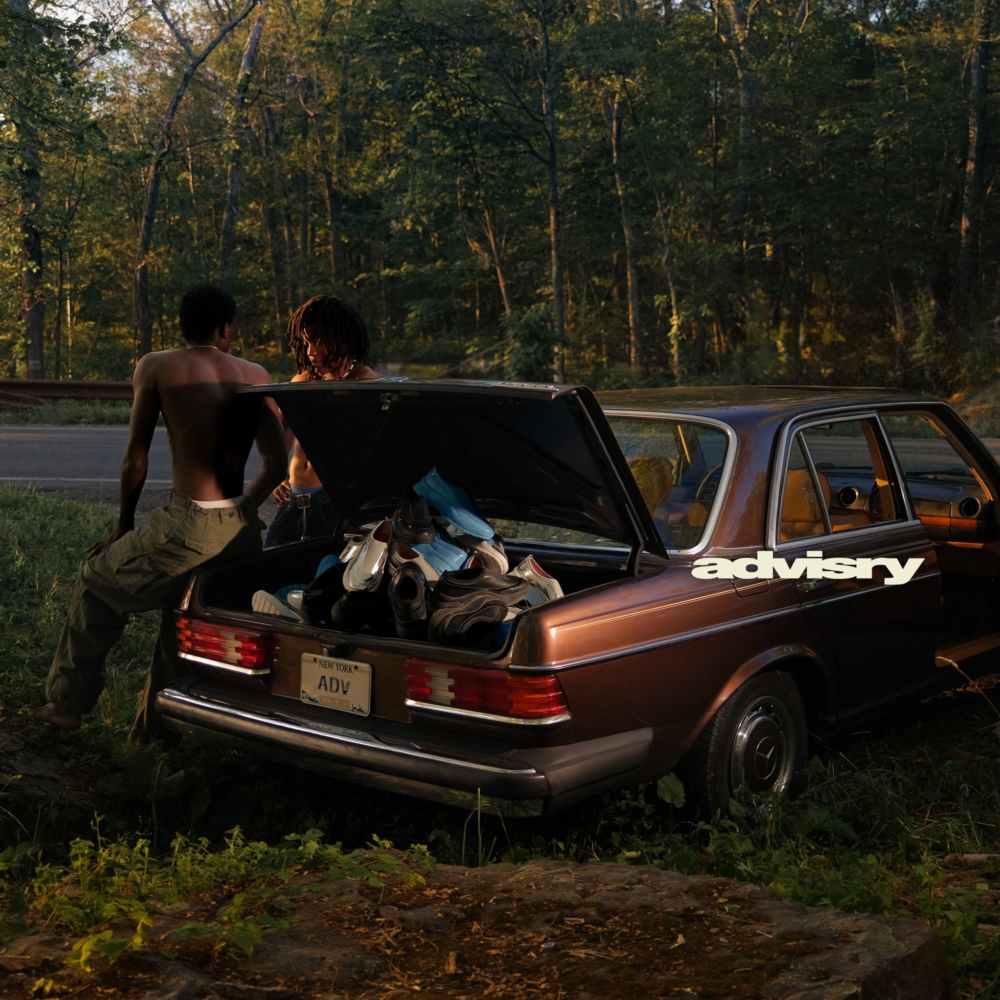 Two shirtless young men beside a brown vintage car with an open trunk piled with sneakers along a wooded road.