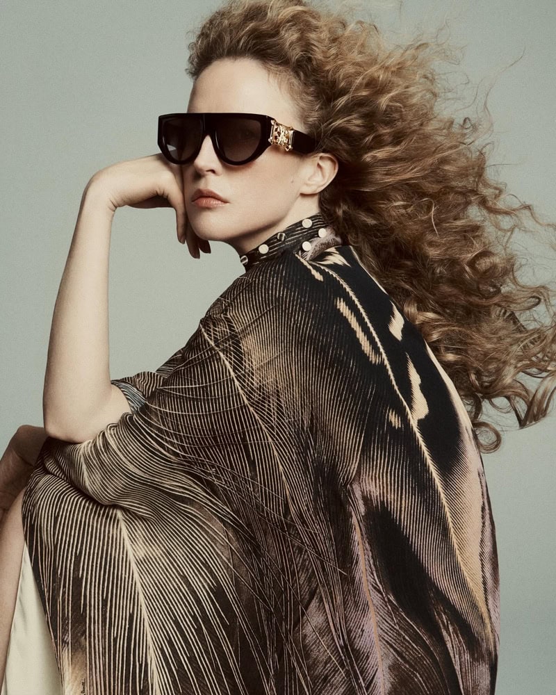 Model wearing dark sunglasses and patterned cape with voluminous curly hair against a grey background