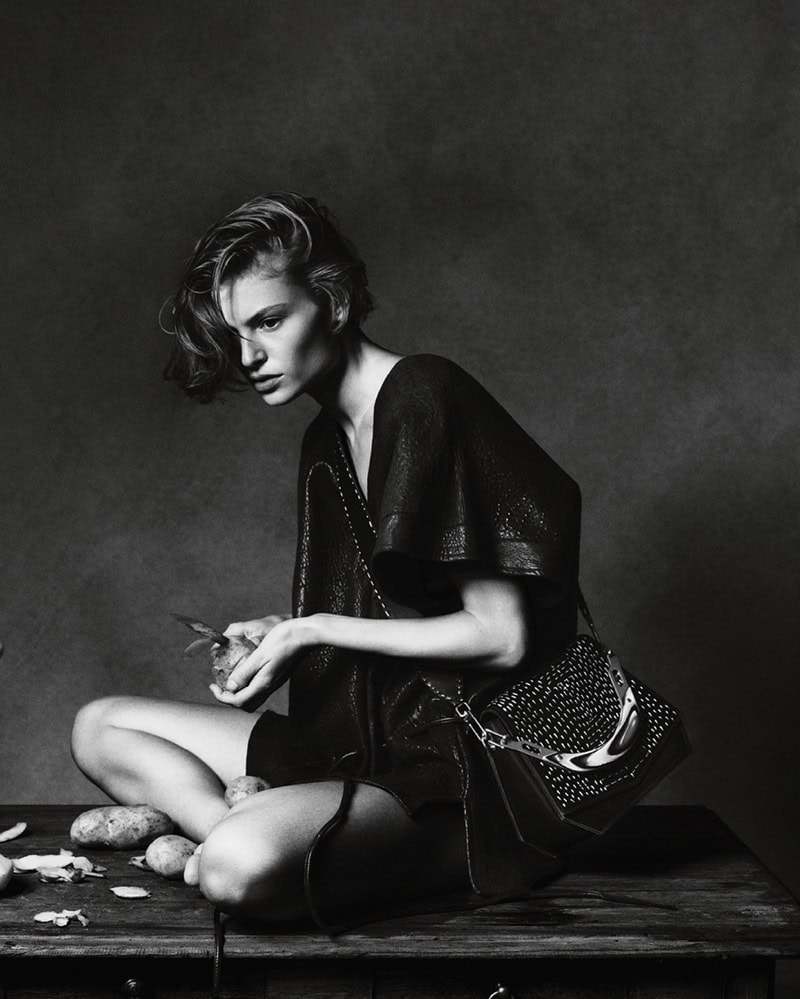 Model seated on a dark wooden table holding a vegetable with a dark shoulder bag against a moody textured backdrop.