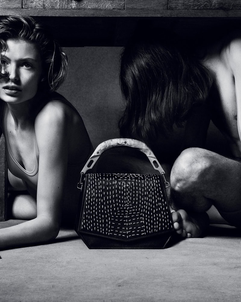 Black and white fashion campaign image featuring a structured handbag with a curved handle and light stitching, positioned before two seated people.