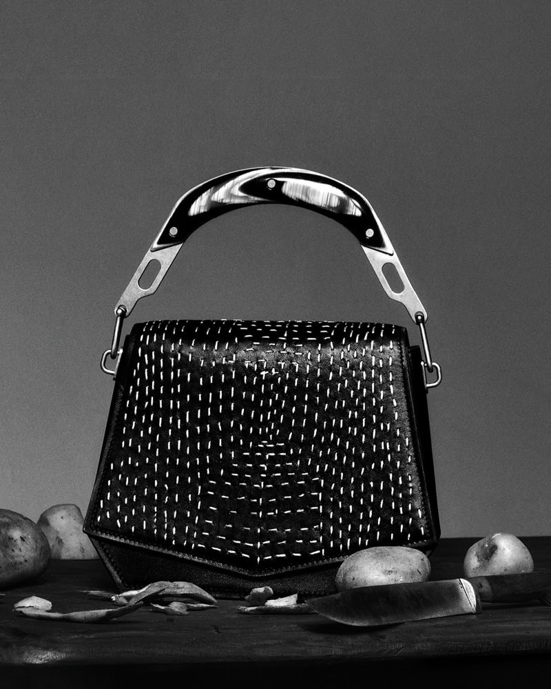 Black handbag with white kantha stitching and a curved handle resting on a dark surface alongside potatoes and a knife.