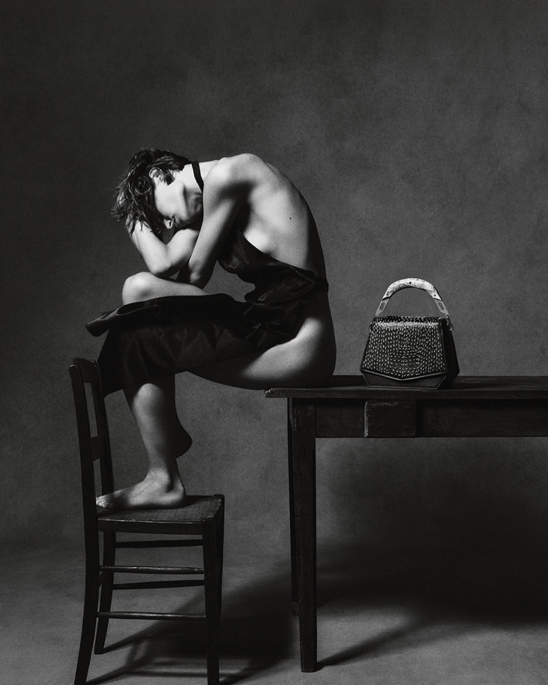 Model seated on a wooden table with one foot on a chair, next to a structured handbag, shot in monochrome.